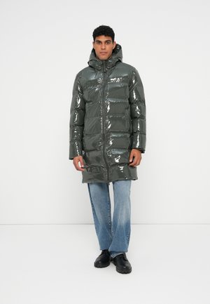 Glänzender, dunkelgrüner Puffer-Mantel mit Kapuze; verfügt über einen Reißverschluss und ein horizontales Steppmuster. Kombiniert mit hellblauen Jeans und schwarzen Stiefeln.