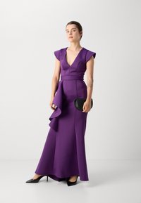 Violet vakarinė suknelė su rankovėmis, giliu V formos iškirpimu ir asimetriška drabužio detalė. Derinama su mažu juodu rankiniu krepšiu ir juodais aukštakulniais batais.