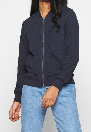 Kvinde iført en marineblå tekstureret lynlåstrøje med hævede prikker og lyseblå denimjeans, stående mod en ensartet hvid baggrund.