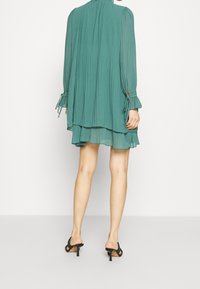 Turkusowa sukienka plisowana z długimi rękawami, akcentami w postaci kokardy, warstwową spódnicą oraz wysokim dekoltem, zestawiona z czarnymi sandałami na obcasie.