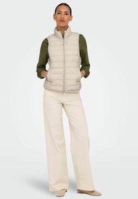Gilet matelassé beige clair avec un col montant et une fermeture éclair à l'avant. Assorti à une blouse verte et un pantalon large crème. Chaussures texturées.