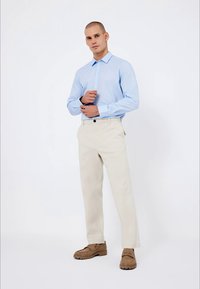 Chemise à manches longues bleu clair avec un col, assortie à un pantalon droit crème et des chaussures brunes à enfiler. Fond uni.