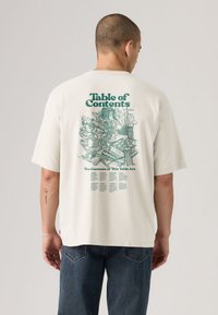 T-shirt de algodão cor creme com estampa gráfica nas costas com o texto "Índice" e ilustrações de vários legumes.
