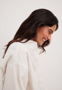 Striped, button-up shirt in cream and light beige fabric. Features a collar and relaxed fit with rolled sleeves, highlighting soft texture.