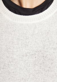 Gros plan sur un cou portant un pull en maille blanc superposé sur un t-shirt noir à col rond avec une texture tricotée visible.