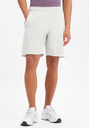 Mann trägt weiße gerippte Shorts, weiße Sneaker und ein lila Hemd, steht mit einer Hand in der Tasche vor weißem Hintergrund.