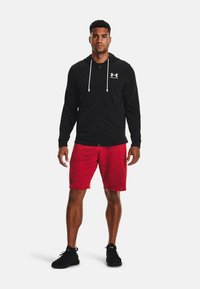 Sort zip-up hoodie med hvide snore, båret over røde pletmønstrede shorts. Sorte atletiske sko fuldender outfittet.