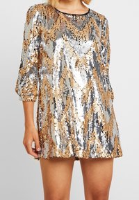 Flitrové šaty s geometrickým cikcak vzorem ve zlaté, stříbrné a černé barvě. Krátké rukávy a kulatý výstřih, zúžené do délky ke středu stehen.