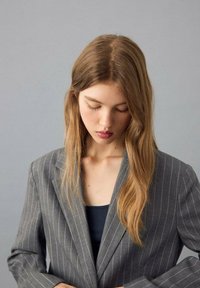 Grijze blazer met krijtstrepen en een gestructureerd ontwerp, voorzien van een revers met inkeping. Lichtgewicht stof met subtiele textuur, gedragen over een donker shirt.