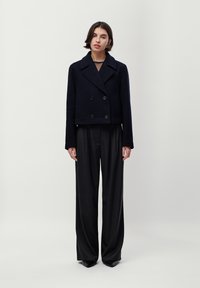 Jachetă scurtă, bleumarin, cu două rânduri de nasturi și guler ascuțit, dintr-un amestec de lână, purtată cu pantaloni largi de culoare închisă, expusă pe un fundal neutru.