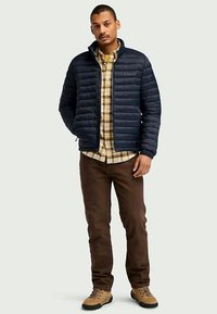 Casaco puffer azul-marinho com costura horizontal, usado sobre uma camisa xadrez amarela e castanha, combinado com calças castanhas e botas bege.