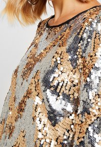 Tílko zdobené flitry s geometrickým vzorem ve zlatých a stříbrných odstínech, s černým výstřihem, měkkou texturou a třpytivým povrchem.