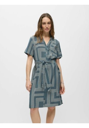 Femme portant une robe à manches courtes avec des boutons, ornée de motifs géométriques bleus et blancs, avec une ceinture nouée à la taille, debout devant un fond clair uni.