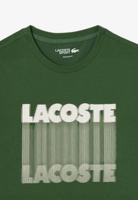 Camiseta de algodón verde con cuello redondo, que presenta un gran logo en degradado blanco de "LACOSTE" en la parte frontal. Diseño de ajuste regular.