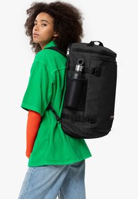 Mochila negra de forma cilíndrica, con un bolsillo de malla en el lateral para una botella de agua, correas ajustables y cremalleras, combinada con una camiseta verde.