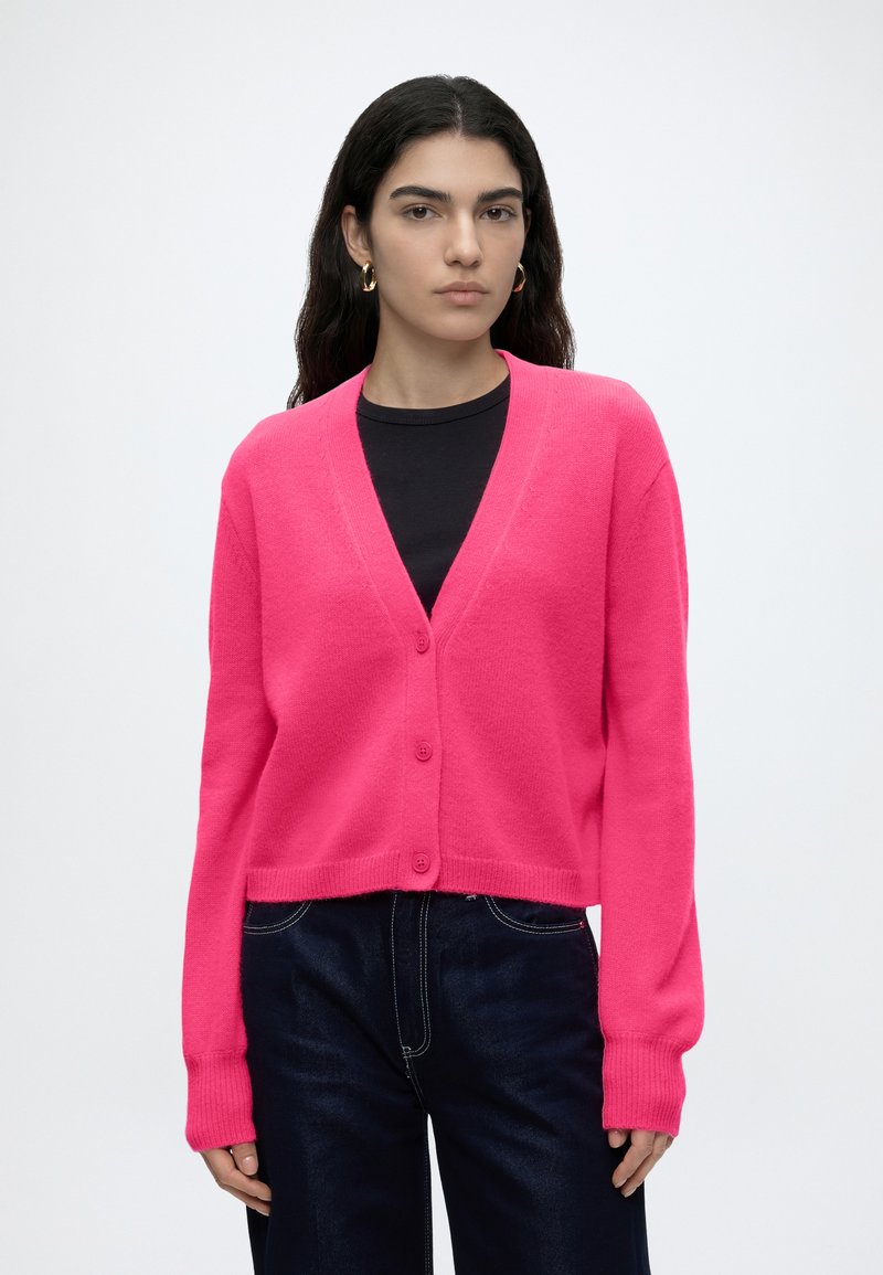 Femme aux longs cheveux foncés porte un cardigan rose vif boutonné par-dessus un top noir et un jean foncé sur un fond clair uni.