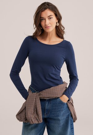 Jeune femme aux cheveux bruns ondulés portant des boucles d'oreilles créoles dorées, une chemise bleu marine à manches longues, un jean bleu et une chemise marron à carreaux nouée autour de la taille.