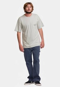 Camiseta de algodón gris claro con un bolsillo en el pecho; combinada con jeans azul oscuro y zapatillas negras con detalles blancos. Ajuste casual, mangas cortas.