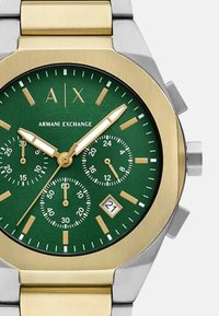 Reloj cronógrafo con una esfera verde, caja de metal dorado y plateado, tres subesferas, indicador de fecha y acentos dorados en los marcadores de horas y en las agujas.