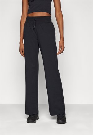 Personne portant un pantalon noir ample, un crop top noir et des baskets noires, debout devant un fond clair uni.