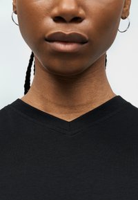 Lähivõte inimese alumisest näost ja kaelast, kes kannab musta V-kaelusega särgikest ja hõbedaseid rõngakõrvarõngaid, nähtavad on punutised juustes.