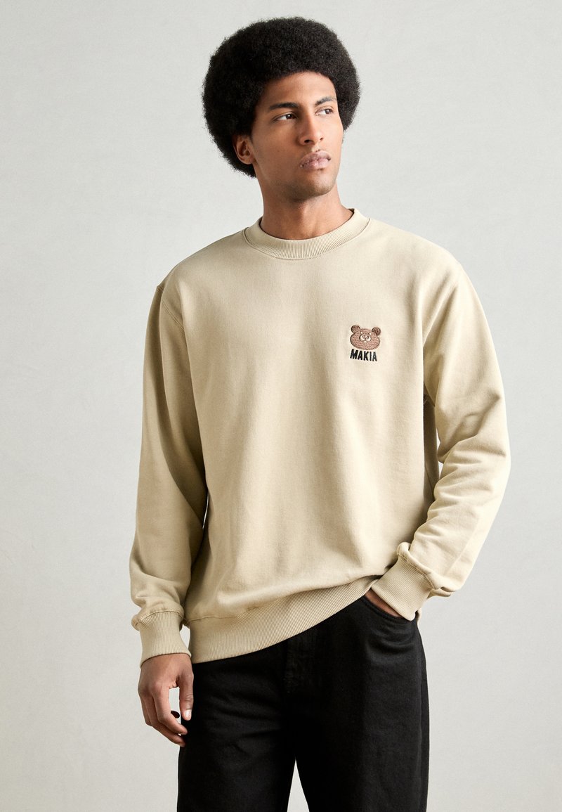 Felpa beige in cotone con ricamo di un orso e logo "MAKIA" sul petto, con polsini e orlo a coste. Pantaloni neri visibili.