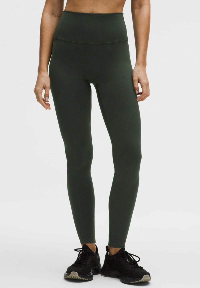 Inimene, kes kannab kõrge vöökohaga tumerohelisi retuuse ja musti spordijalatseid, seisab tavalise heleda tausta ees.