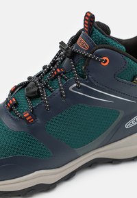 Wasserfeste Wanderschuhe in dunklem Teal und Navy, mit einem strukturierten Mesh-Obermaterial, verstellbaren Schnürsenkeln und einer robusten grauen Sohle mit Grip.