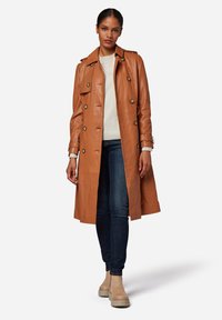Tan leather trench coat with double-breasted buttons, shoulder epaulettes, and a belted waist. Paired with blue jeans and beige ankle boots.