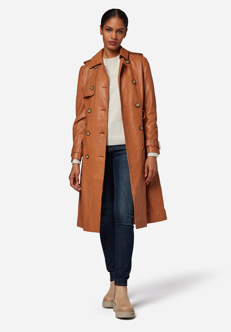Tan leather trench coat with double-breasted buttons, shoulder epaulettes, and a belted waist. Paired with blue jeans and beige ankle boots.