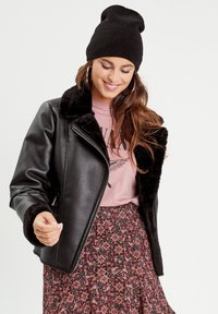 Veste en simili cuir noir avec col en fausse fourrure, devant zippé et poignets épais, associée à une jupe rose à motifs et un bonnet noir.