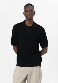 Μαύρο πόλο μπλουζάκι κοντό μανίκι φτιαγμένο από ριπ ύφασμα. Διαθέτει κλασικό γιακά και τρεις κουμπιά. Φοράει με ανοιχτόχρωμο παντελόνι.