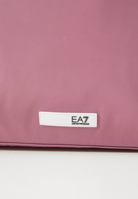 Rosa syntetisk veske med en glatt tekstur. Har en hvit etikett med "EA7 EMPORIO ARMANI" logo. Enkel, rektangulær design.
