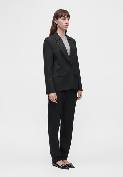 Femme debout portant un costume noir sur mesure avec un blazer à un seul bouton, un pantalon noir, un haut gris et des chaussures plates pointues noires.
