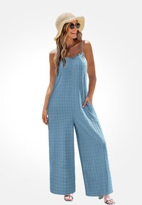 Vrouw die glimlacht in een lichtblauwe wijde jumpsuit met zakken, draagt een zonnehoed, ronde zonnebril en gelaagde gouden kettingen.