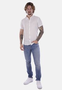 Camicia bianca in lino a maniche corte con bottoni, abbinata a jeans in denim blu chiaro e sneakers bianche. Presenta tatuaggi visibili sulle braccia.