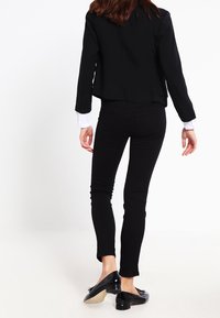 Musta räätälöity bleiseri valkoisen paidan päällä, yhdistettynä vartalonmyötäisiin mustiin farkkuihin ja mustiin loafereihin. Suoria pintakäsittelyitä, lyhennetty malli.