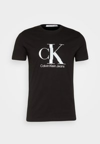 Černé bavlněné tričko s velkým bílým logem "CK" a textem "Calvin Klein Jeans" pod ním. Krátké rukávy a kulatý výstřih.