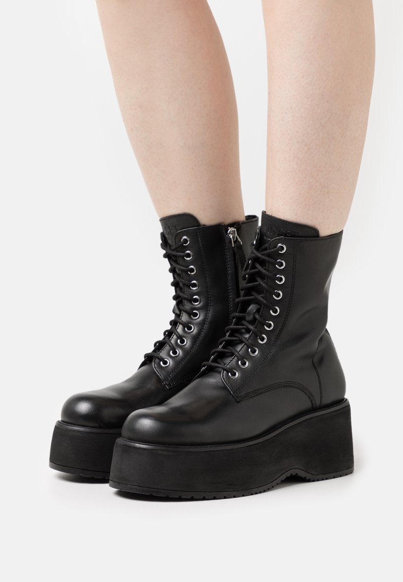 Botas de tobillo de cuero negro con punta redonda, cierre de cordones en la parte delantera, cremallera lateral y suela gruesa de plataforma. Textura suave con acentos mínimos.