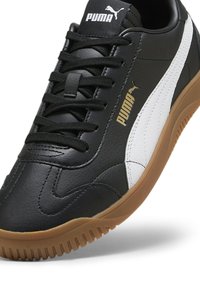 Zapatilla negra de cuero sintético de Puma con franja lateral blanca, logo dorado, cordones negros y suela de goma marrón. Diseño bajo, parte superior texturizada.