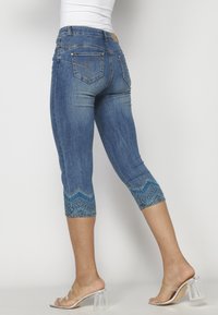 Femme portant un jean en denim bleu court avec un détail brodé bleu sur les ourlets et des sandales à talons transparents, vue de dos.