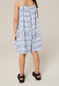 Dziewczyna w bezrękawnikowej niebiesko-białej sukience w wzory, w czarnych sandałach, stojąca na jasnym jednolitym tle, odwrócona plecami.