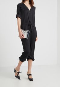 Combinaison noire à manches courtes, avec fermeture éclair à l'avant et taille élastique. Associée à des chaussures à talons hauts noires, avec un accent nœud et une pochette argent.