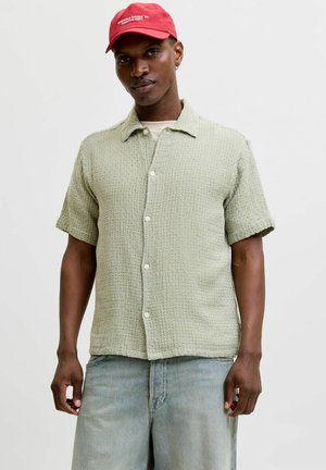 Homme portant une chemise à manches courtes boutonnée de couleur vert clair texturée, un jean bleu clair et une casquette rouge avec un texte blanc sur un fond uni.