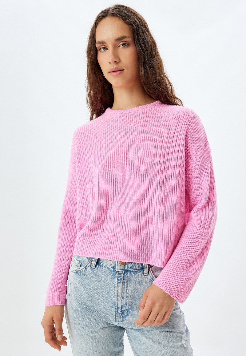 Rosa stickad tröja med rund halsringning, ribbad struktur och sänkta axlar, bärs över ljusblå slitna jeans.