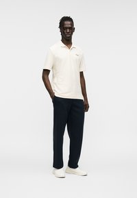 Ανοιχτό μπεζ πόλο μπλουζάκι με διακριτική ριγέ υφή συνδυασμένο με σκούρο μπλε παντελόνι. Το μοντέλο φορά λευκά αθλητικά παπούτσια και στέκεται μπροστά σε απλό φόντο.