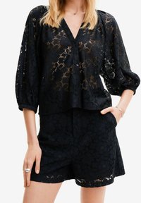 Una modella che indossa un top di pizzo nero con scollatura a V e maniche sciolte, abbinato a pantaloni corti coordinati, che presentano motivi floreali.