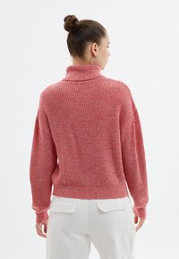 Pull en laine rouge tricoté avec un col montant, coupe large et texture côtelée. Présente des épaules tombantes et une couleur unie avec de légères variations.