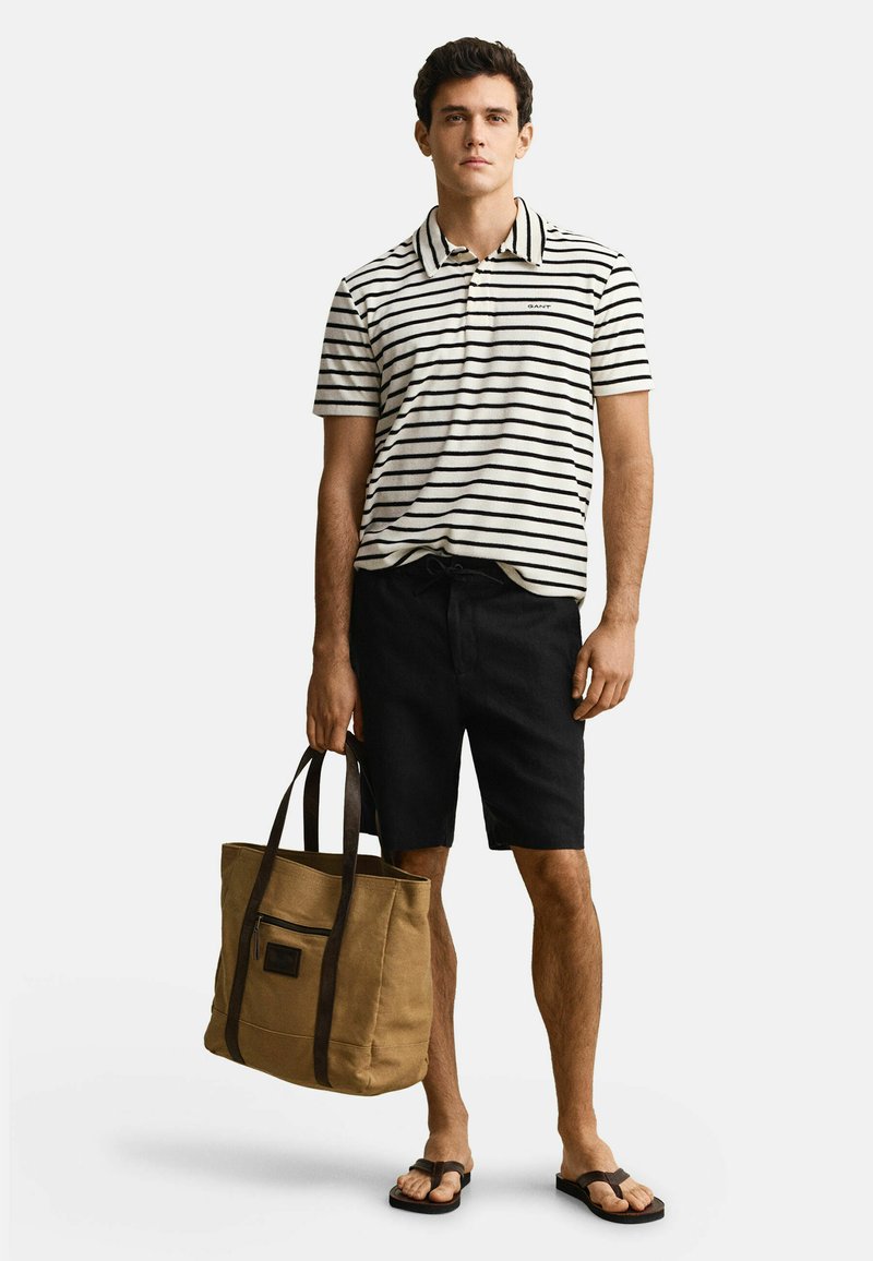 Homme portant un short noir, un polo à manches courtes rayé, des tongs, tenant un grand sac fourre-tout marron avec des poignées foncées.