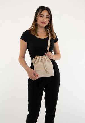 Beige bucket bag made of textured leather with gold hardware. Drawstring closure and adjustable strap. Worn by a person in black attire.
